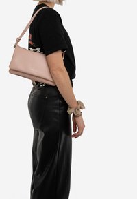 Jasnoróżowa skórzana torba na ramię z węzłowym paskiem, o gładkiej teksturze i minimalistycznym designie. Noszona z czarnymi skórzanymi spodniami.