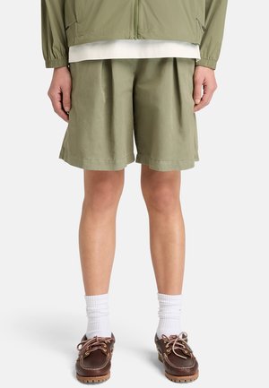 Persona che indossa pantaloncini plissettati verde oliva chiaro, calzini bianchi e scarpe da barca di cuoio marrone, in piedi contro uno sfondo semplice.