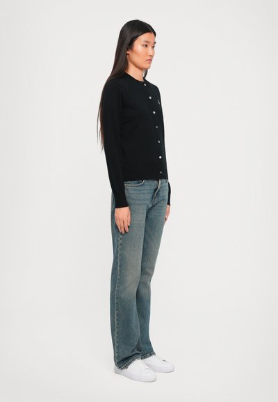 Donna in piedi di lato, che indossa una cardigan nera con bottoni, jeans blu a gamba dritta e sneakers bianche, su uno sfondo semplice.