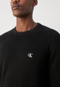 Melns adīts džemperis ar apaļu kakla izgriezumu un baltu izšūtu "CK" logo kreisajā krūtīs. Gluda tekstūra visā virsmā.