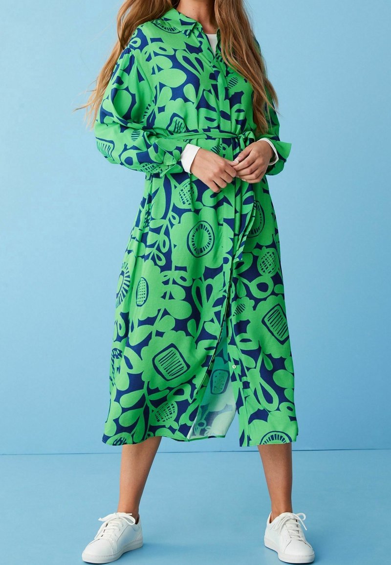 Femme portant une robe midi à manches longues à fleurs vertes et bleues avec ceinture et baskets blanches sur fond bleu clair.