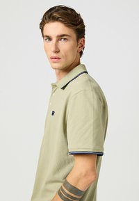 Piké i ljusgrön bomull med en texturerad yta, krage med marinblå detaljer och en liten logotyp på bröstet.