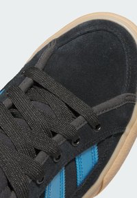 Čierna semišová teniska s modrými akcentmi. Má ploché čierne šnúrky, textúrovaný zvršok a svetlú gumovú podrážku.