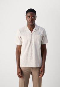 Camisa de manga corta, texturizada en beige claro, con botón en la parte frontal y cuello abierto. Presenta dos bolsillos en el pecho y un ajuste relajado.