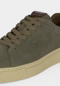 Baskets en daim vert olive avec lacets beige, semelle beige et languette de talon en cuir marron, avec le logo "GANT" sur le côté et la languette.