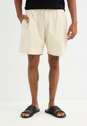 Hombre con pantalones cortos beige con cordón y bolsillos, y sandalias negras tipo slide, de pie contra un fondo blanco liso.