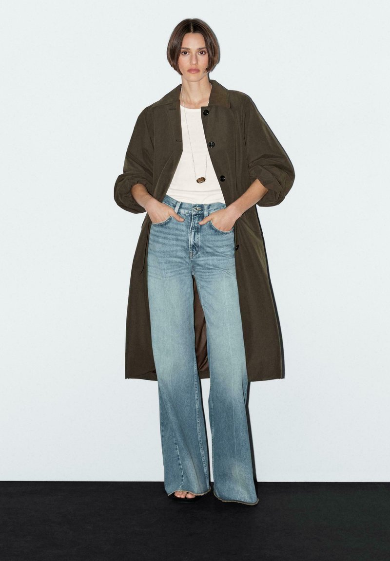 Trench coat verde oliva con maniche a palloncino, abbinato a jeans a zampa di elefante blu chiaro. T-shirt bianca sotto, collana con pendente minimalista.