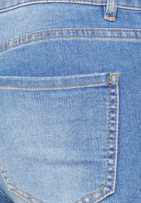 Jeans de ganga azul claro com um único bolso traseiro. Apresenta costura reta em fio laranja contrastante e um acabamento suave e texturizado.
