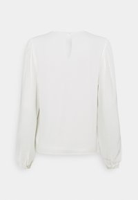 Blouse blanche à manches longues, en tissu doux, avec un col arrondi et une petite ouverture en forme de clef dans le dos. Présente des détails froncés au niveau des épaules.