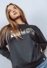 Młoda kobieta o długich blond włosach, ubrana w czarną bluzę z odblaskowymi napisami, spoglądająca lekko w górę na jednolitym tle.