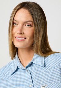 Niebiesko-biała koszula z guzikami w kratę, z kołnierzykiem, wyposażona w kieszeń oraz subtelną metkę z logo. Gładki bawełniany materiał o luźnym kroju.