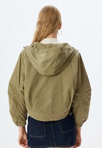 Olivengrøn, letvægtsjakke med hætte, elastiske manchetter og en cropped silhuet. Glat tekstur, har sidelommer og lynlåse.