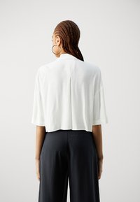 Camicia bianca cropped con maniche corte e dettagli di apertura, abbinata a pantaloni neri a gamba larga. La trama sembra leggera e liscia.