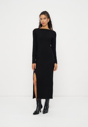 Femme aux cheveux bouclés portant une robe noire à manches longues avec une fente haute sur le côté et des bottines noires sur un fond blanc uni.