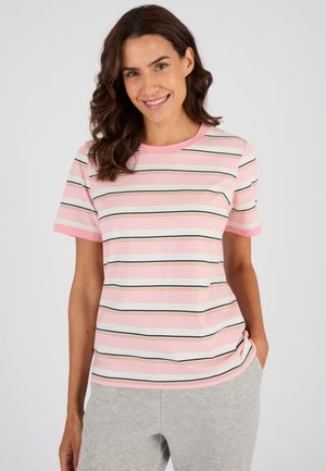 Donna sorridente con capelli castano ondulati, indossa una maglietta a maniche corte a righe rosa e bianche e pantaloni grigi chiari, in piedi con le mani nelle tasche.