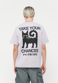 Lyse lilla bomulds t-shirt med et sort kattemotiv og teksten "TAG DINE CHANCER" på ryggen; korte ærmer og afslappet pasform.