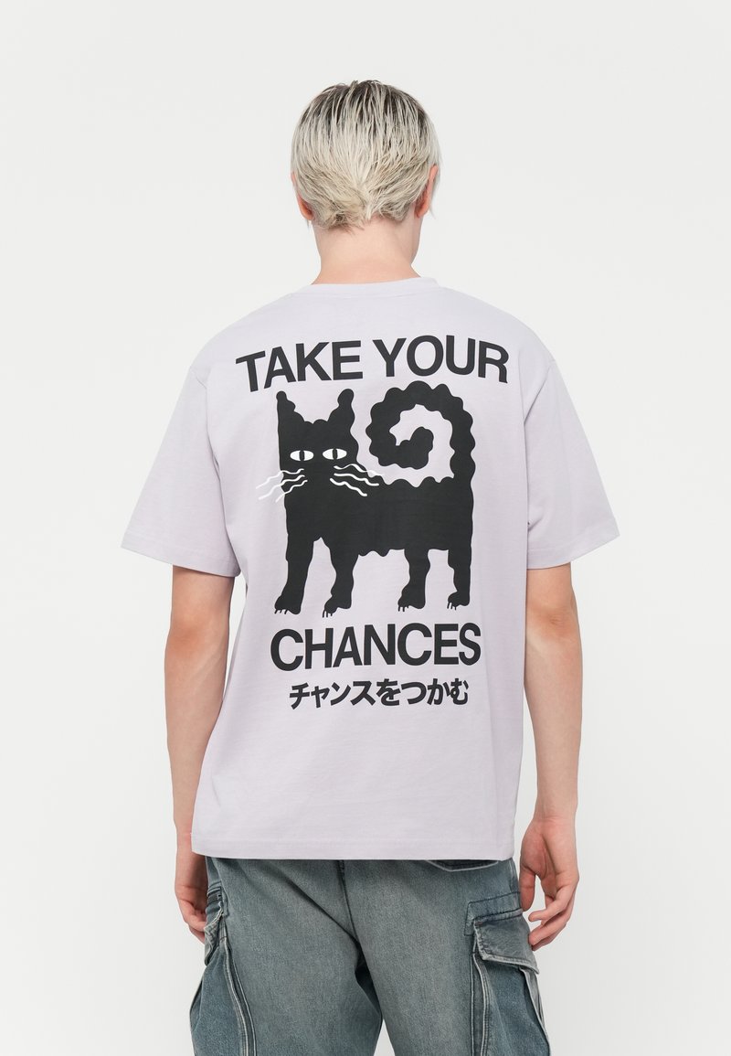 Lyse lilla bomulds t-shirt med et sort kattemotiv og teksten "TAG DINE CHANCER" på ryggen; korte ærmer og afslappet pasform.