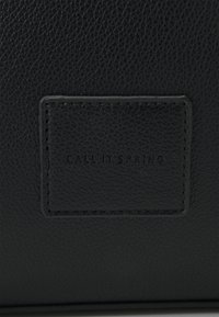 Sac en cuir texturé noir avec une étiquette rectangulaire embossée de "CALL IT SPRING", soulignée d'un surjet contrastant.