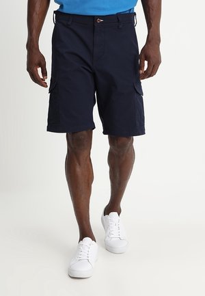 Man draagt donkermarineblauwe cargoshorts en witte sneakers en loopt voor een effen lichte achtergrond.