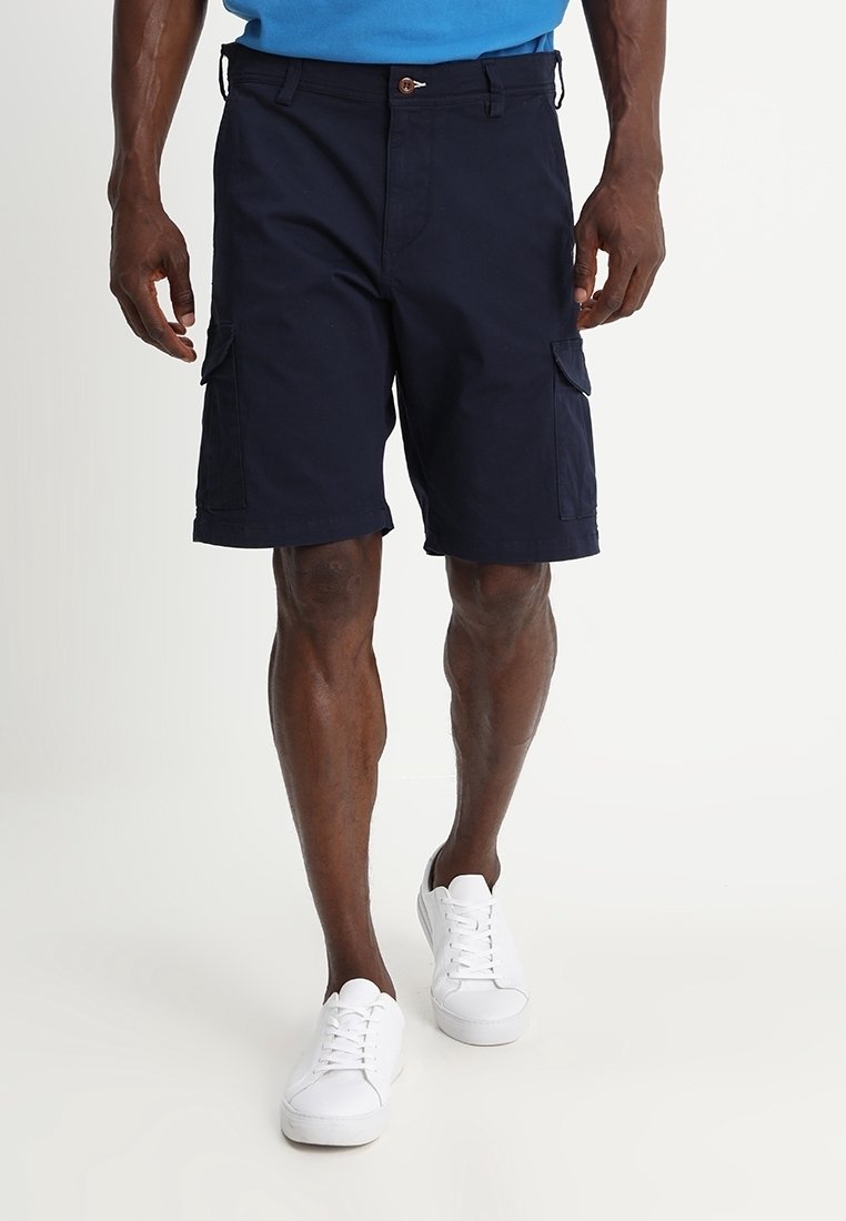 Homme portant un short cargo bleu marine foncé et des baskets blanches marchant devant un fond clair uni.