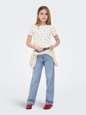 Ragazza con capelli lunghi che indossa una camicia bianca con puntini rossi, jeans azzurri chiari, scarpe rosse e un maglione beige legato in vita, in piedi e rivolta verso di noi.