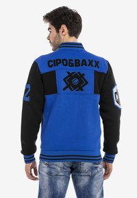 Blaue und schwarze Varsity-Jacke mit gestreiften Bündchen und Kragen. Mit gestickten Logos und Zahlen auf dem Rücken und dem Ärmeln.