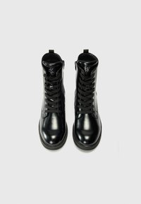 Bottes cheville à lacets en cuir verni noir avec bouts arrondis et logo Calvin Klein sur la languette, vues de dessus sur fond blanc.