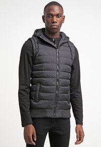 Vest negru căptușit cu glugă, cu fermoar în fa�ță și buzunare laterale, având un model pătrățel orizontal și tiv zimțat. Purtați peste un tricou negru cu mânecă lungă.
