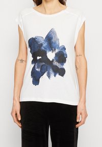 Camiseta de manga corta blanca, fabricada en suave algodón, con un gran gráfico floral azul impreso en la parte frontal. Diseño sencillo y de corte relajado.