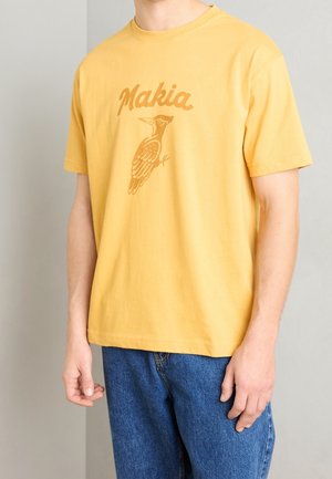 Person trägt ein gelbes T-Shirt mit kurzen Ärmeln mit "Makia" und einer stilisierten Vogeldarstellung, kombiniert mit blauen Jeans.