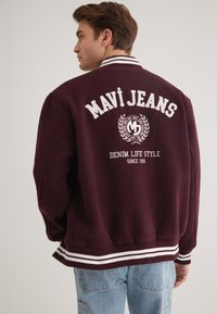 Bordeauxfarbener Varsity-Jacket mit weißer Schrift, ausgestattet mit einem Rippkragen und -bündchen sowie einem Logo mit Lorbeerdesign auf dem Rücken. Lässiger Schnitt, aus Fleece-Material.