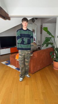 Νέος άνδρας με πουλόβερ με ρίγες και παντελόνι παραλλαγής στέκεται με σταυρωμένα τα χέρια σε ένα μοντέρνο σαλόνι δίπλα σε καφέ καναπέ και μεγάλο φυτό.