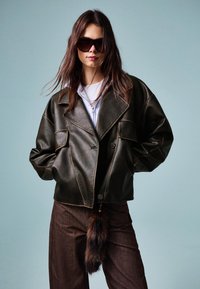 Hnedá falošná kožená bunda so širokým golierom, veľkými vreckami a kontrastným šitím, skombinovaná s hnedými nohavicami s akcentom z falošnej kožušiny.