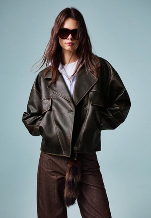 Giacca in finta pelle marrone con colletto largo, ampie tasche e cuciture a contrasto, abbinata a pantaloni marroni con un accento in pelliccia sintentica.