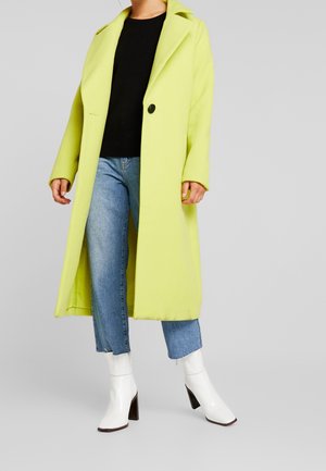 Persona que lleva un abrigo largo amarillo brillante, camiseta negra, vaqueros azules y botines blancos con tacón, de pie frente a un fondo sencillo.