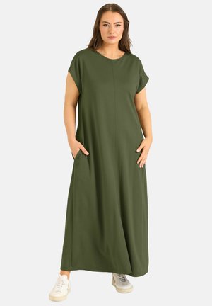Kobieta stojąca z rękami w kieszeniach, ubrana w oliwkowozieloną, krótkorękawową sukienkę maxi i białe trampki na białym tle.
