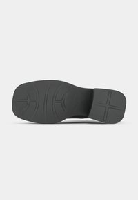 Semelle de chaussure noire avec des motifs texturés et rainurés, comprenant des lignes courbes et une forme de croix au niveau du talon, présentée sur un fond uni.