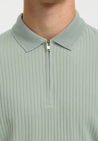 Gros plan d'une personne portant un polo côtelé vert clair avec un col texturé et une fermeture éclair partiellement fermée au niveau du cou.