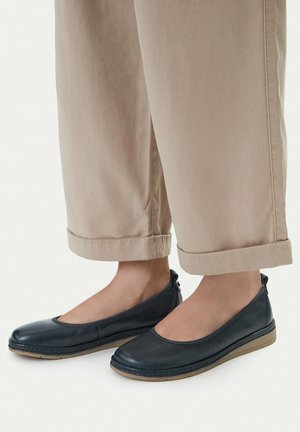 Persona con pantalones beige arremangados y zapatos planos de cuero negro sin cordones con suelas color marrón claro, mostrado desde el tobillo hacia abajo sobre un fondo blanco.