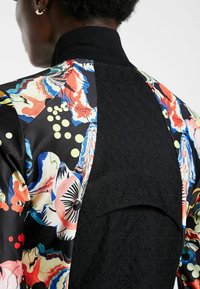 Kolorowa kurtka bomberka z kwiatowym wzorem z jednej strony i teksturowanym czarnym materiałem z drugiej, z prążkowanym kołnierzem i zapięciem na guziki.
