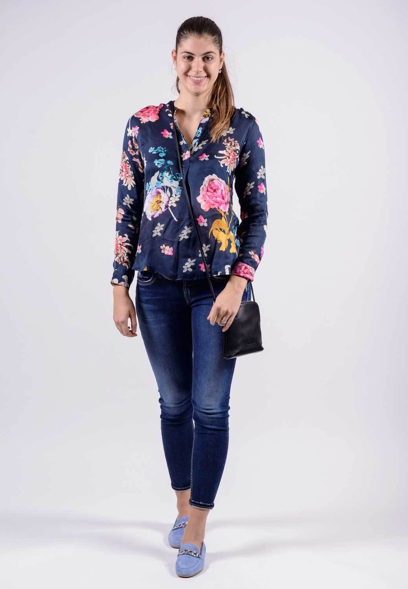 Blaues Blumenblusenoberteil mit Knöpfen, taillierte Form, kombiniert mit blauen Jeans und pastellfarbenen Slippern. Schwarze Umhängetasche mit dünnem Riemen.