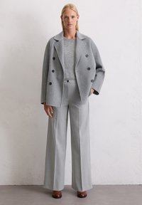 Donna che indossa un cappotto grigio doppiopetto, un maglione di maglia grigia abbinato, pantaloni a gamba larga e scarpe marroni, in piedi contro un muro bianco testurizzato.