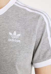 Šedé bavlněné tričko s bílým ribbovaným výstřihem a pruhy na ramenou. Na hrudi je bílé vyšívané logo Adidas.