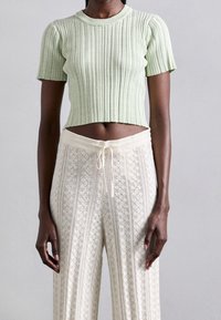 Osoba o ciemnej karnacji nosząca jasnoszary prążkowany, krótki top z krótkim rękawem oraz białe, szydełkowe spodnie z wysokim stanem i sznurkiem.