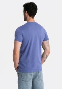 T-shirt a maniche corte e collo rotondo in morbido cotone viola, indossato con jeans in denim azzurro chiaro. Tatuaggi visibili sul braccio.