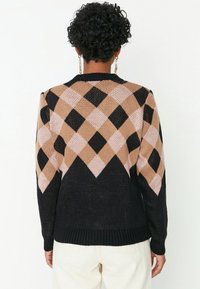 Personne aux cheveux courts et bouclés portant un pull à motif losanges noir, beige et rose clair ainsi qu'un pantalon de couleur claire, vue de dos.
