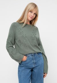 Pull en tricot vert avec broderies florales, manches larges et poignets côtelés. Associé à un jean bleu taille haute. Texture douce.