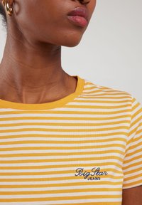 Dzeltena un balta svītraina T-krekls ar apaļu kaklu un izšūtu "Big Star Jeans" logotipu kreisajā pusē. Gluda kokvilnas tekstūra.