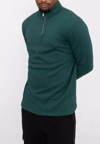 Pull en maille vert avec col montant zippé. Le tissu présente un motif de diamant texturé, avec des manches longues et une silhouette ajustée.