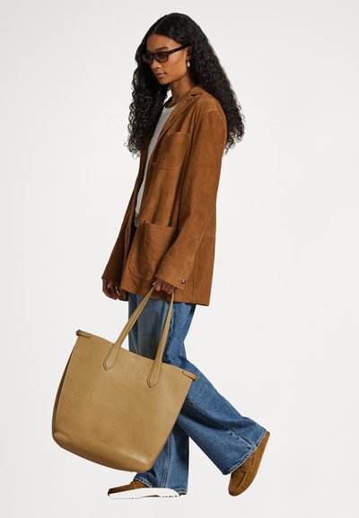 Torba iz rjavkastega usnja s dvema dolgima ročajema, ima gladko teksturo in prostorno obliko. Nošena z rjavim antilop blazerjem in modrimi jeans hlačami.